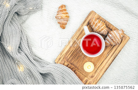 A cup of tea, a candle, a croissant and a knitted element in bed, top view. 135875562