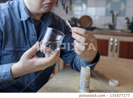 A man in the kitchen tests tap water quality with a test strip. Home testing for drinking water safety, pH, chlorine, and chemical contaminants. A man in the kitchen tests tap water quality with a test strip. Home testing for drinking water safety, pH, chlorine, and chemical contaminants. 135877177