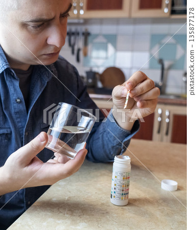 A man in the kitchen tests tap water quality with a test strip. Home testing for drinking water safety, pH, chlorine, and chemical contaminants. A man in the kitchen tests tap water quality with a test strip. Home testing for drinking water safety, pH, chlorine, and chemical contaminants. 135877178