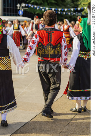 Folk dancers in traditional Bulgarian costumes performing in public square. People in in folk costumes holding hands dancing in circle at festival. Colorful national outfits with intricate embroidery 135877790