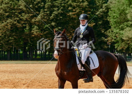 A female hijab equestrian athlete on horseback is holding a gold medal after winning a dressage competition. This image represents achievement, success, and dedication in sports, showcasing the 135878102