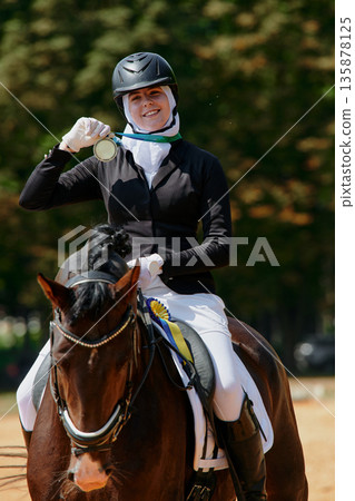 A female hijab equestrian athlete on horseback is holding a gold medal after winning a dressage competition. This image represents achievement, success, and dedication in sports, showcasing the 135878125
