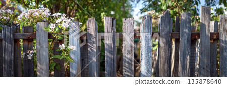 A fence made from recycled wooden planks and metal scraps encloses a suburban backyard. Green plants and flowers grow around the fence on a sunny day, banner A fence made from recycled wooden planks and metal scraps encloses a suburban backyard. Green plants and flowers grow around the fence on a sunny day, banner 135878640