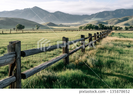 A weathered wooden farm gate stands in front of lush green pasture, surrounded by tall grass and distant hills under a cloudy sky, showing a serene rural scene 135878652