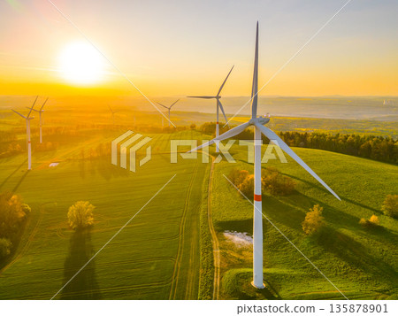 Wind turbines are rotating in a field during sunset. The sun casts a bright light over the landscape. Trees are visible nearby as shadows stretch across the ground. Wind turbines are rotating in a field during sunset. The sun casts a bright light over the landscape. Trees are visible nearby as shadows stretch across the ground. 135878901