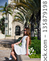 Woman in white dress sitting by palm trees and historic building in sunlight 135879198