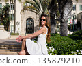 Smiling woman in white dress sitting outside with palm trees and city background 135879210