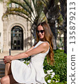Stylish woman in white dress sitting outdoors by palm trees and stone building 135879213