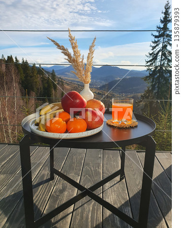 composition of fruits on a plate: bananas, apples, oranges, juice in a glass, a vase with dry plants on a black metal table on a wooden terrace with a view of the Carpathian Mountains. Majestic peaks 135879393