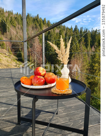 composition of fruits on a plate: bananas, apples, oranges, juice in a glass, a vase with dry plants on a black metal table on a wooden terrace with a view of the Carpathian Mountains. Majestic peaks 135879492