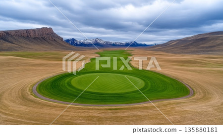 A lush green golf course fairway surrounded by dry fields and distant mountains under cloudy skies 135880181