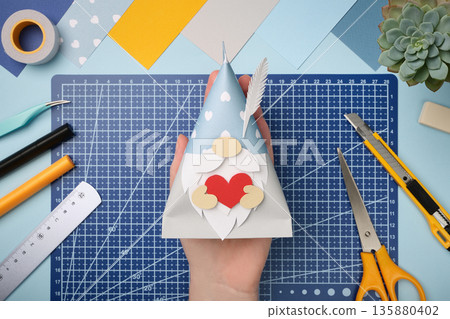 Craft made of colored paper. Gnome with heart in hand. Creative hobby and master class idea. Craft made of colored paper. Gnome with heart in hand. Creative hobby and master class idea. 135880402