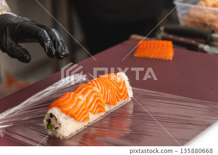 Chef in black glove preparing fresh salmon sushi roll Chef in black glove preparing fresh salmon sushi roll 135880668
