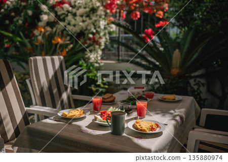 Outdoor breakfast with dishes and drinks on a table surrounded by flowers and plants 135880974
