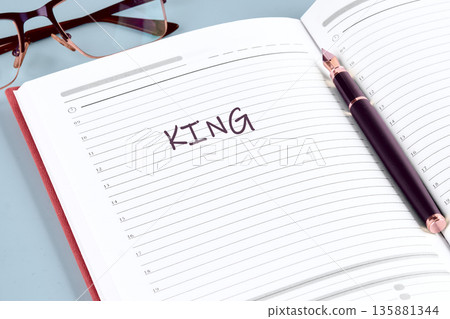 A neat desk setup shows an open daily planner with the word KING written in black ink, resting beside a sophisticated dark fountain pen and a pair of reading eyeglasses A neat desk setup shows an open daily planner with the word KING written in black ink, resting beside a sophisticated dark fountain pen and a pair of reading eyeglasses 135881344