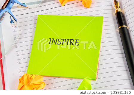 Green sticky note displays Inspire text, centrally placed on an open lined planner. Blue reading glasses and a black fountain pen rest close Green sticky note displays Inspire text, centrally placed on an open lined planner. Blue reading glasses and a black fountain pen rest close 135881350