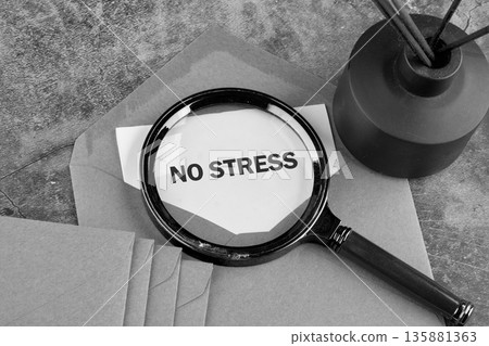 A magnifying glass reveals the phrase NO STRESS on a white paper. Several brown envelopes are visible below. A dark diffuser with reeds stands on the desk surface 135881363