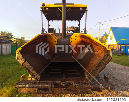 Orange Road Construction Paving Machine Without Driver Standing on Road with Stone Compacted Base. Empty Container, Front View. Replacement floors with modern materials, durability, bearing capacity Orange Road Construction Paving Machine Without Driver Standing on Road with Stone Compacted Base. Empty Container, Front View. Replacement floors with modern materials, durability, bearing capacity 135881593