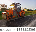 Orange asphalt tamping machine with heavy large vibrating wheels standing parked on side of the road next to new asphalted rural road in private household area. Machines for road construction works 135881596