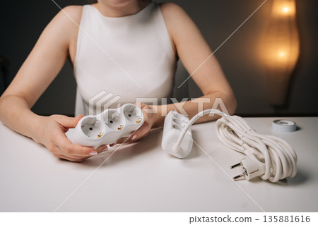Woman holding white triple electrical socket in her hands, sitting at desk with another socket next to her, close-up. Concept of electrical solutions, home improvement. 135881616