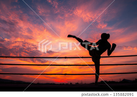 Muay thai boxing ritual performed by female fighter in silhouette with dramatic sunset sky background showing strength and tradition in outdoor ring 135882091