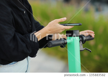 Cropped photo of woman with smartphone scans QR code for electric scooter rental. Girl using app on phone to unlock dockless transport outdoors. Urban riding ecological vehicle. Modern technology 135883582