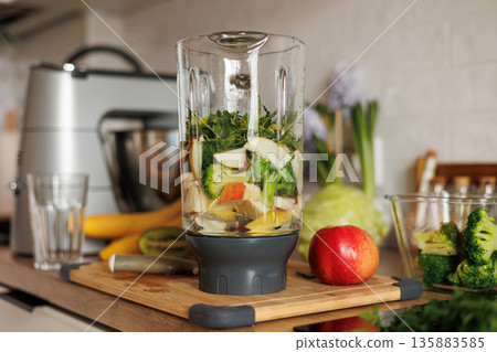 Glass blender bowl with apple, banana and broccoli slices, parsley and water inside on a wooden table on home kitchen. Healthy detox smoothie drink and lifestyle diet. Pieces of fruit ingredients. Glass blender bowl with apple, banana and broccoli slices, parsley and water inside on a wooden table on home kitchen. Healthy detox smoothie drink and lifestyle diet. Pieces of fruit ingredients. 135883585