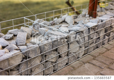 Stone fence in steel mesh and green lawn. Close up of gabion with tightly packed stones of varying sizes and textures. Combination of natural materials and industrial design. 135883587