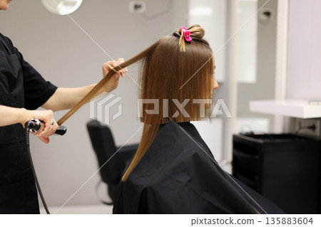 Unrecognisable professional hairdresser ironing female client hair with hair straightener and comb, making stylish hairstyle in beauty salon. Keratin, lamination, hair care, hair salon and hairstyling 135883604