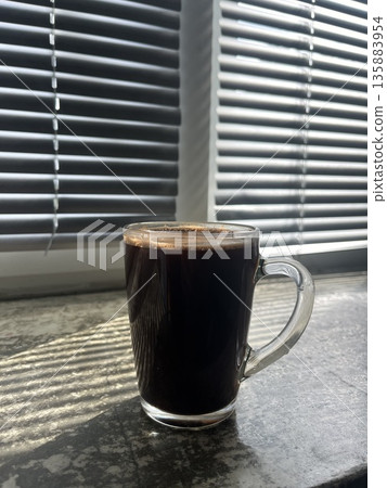 A large cup of coffee on the windowsill near the blinds.  135883954