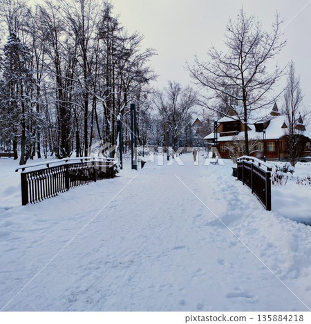 Snow-covered park scene featuring a wooden bridge leading to a charming rustic cabin Snow-covered park scene featuring a wooden bridge leading to a charming rustic cabin 135884218