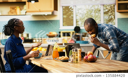 African american man in a hurry running late for his office job, skipping breakfast and leaving his housewife to eat alone at the table. Boyfriend waving goodbye and rushing to leave. 135884420