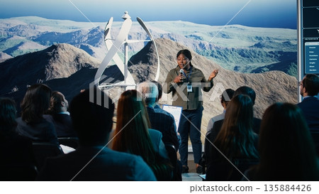 Black female speaker does wind power presentation at business forum with wind turbine machine and rotation. Addressing listeners during public speaking event focused on alternative energy. 135884426