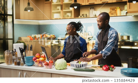 African american people cutting fresh vegetables on a cutting board at home, slicing cucumber and bell pepper for fresh vegetarian salad. Man and woman enjoying meal prep. 135884429