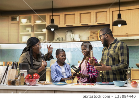 African American family enjoying silly karaoke session for pizza day in the kitchen, singing with utensils and doing goofy dance moves together at home. Weekend activity and cooking time. African American family enjoying silly karaoke session for pizza day in the kitchen, singing with utensils and doing goofy dance moves together at home. Weekend activity and cooking time. 135884440