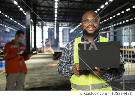 African american supervisor standing near steel machinery with laptop during construction tasks, showcasing production responsibility and the power of fabrication. Hi vis vest for safety. 135884458