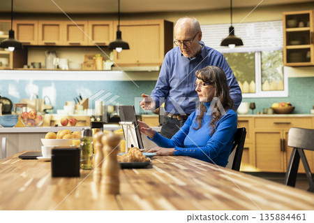 Aged couple checking apps on tablet while they sit together in the kitchen, enjoying fresh coffee drink and casual conversation indoors. Candid moment during a relaxed family morning. Aged couple checking apps on tablet while they sit together in the kitchen, enjoying fresh coffee drink and casual conversation indoors. Candid moment during a relaxed family morning. 135884461