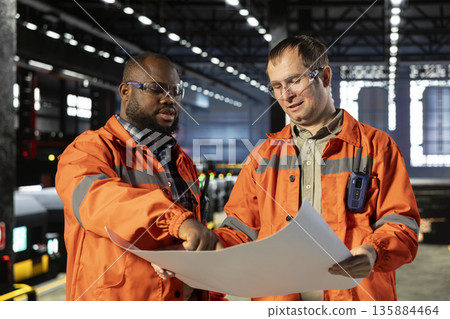 Diverse operators handling assembly workflow with industrial plans, standing near steel machinery and fabrication stations that represent heavy industry development. Welding sparks. Diverse operators handling assembly workflow with industrial plans, standing near steel machinery and fabrication stations that represent heavy industry development. Welding sparks. 135884464