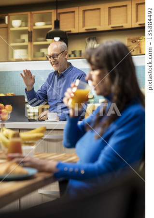 Mature pensioner enjoys happy video conference with family on his laptop. Senior man relaxing with coffee and pastry on the counter, showing a positive online communication during retired life. 135884492
