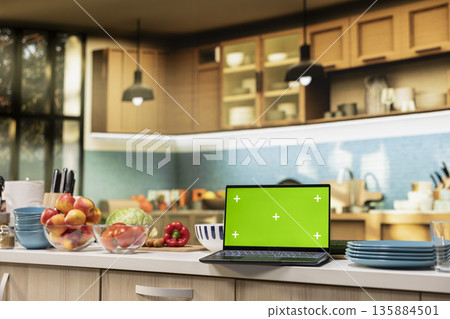 Green screen on laptop in the kitchen with rustic counter and elegant lighting. Cookware and utensils creating a comfortable and inviting household dining space with modern interior design. 135884501