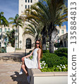 Confident woman in white dress sitting outdoors near palm trees and building 135884813