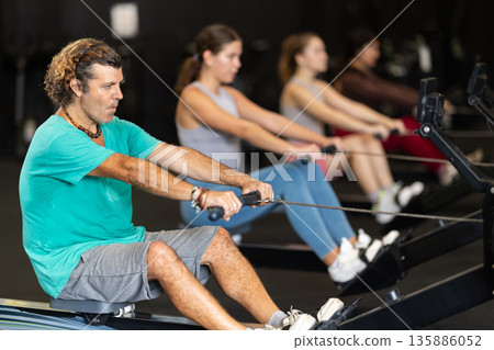Diligent middle-aged man training with rowing machine in sports hall Diligent middle-aged man training with rowing machine in sports hall 135886052