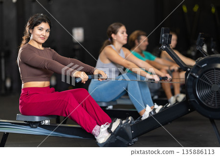 Positive Latina girl training with rowing machine in sports hall 135886113