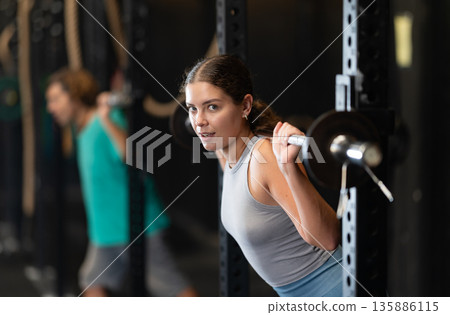 Positive young woman lifting barbell in sports hall 135886115