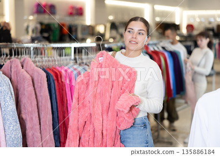 Girl stopped near leisure wear showcase, holds eiderdown bathrobe hanger in hands, examines product Girl stopped near leisure wear showcase, holds eiderdown bathrobe hanger in hands, examines product 135886184