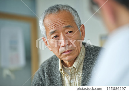 A senior Japanese man receiving consultation and examination from a doctor or specialist. Image of medical care, nursing care, health anxiety and resolution. 135886297