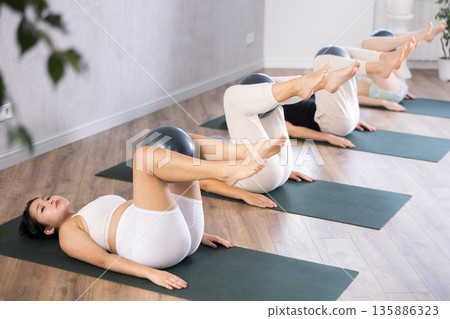 Girl is doing Pilates in the studio. People lie on their backs holding a soft ball pilates in their outstretched legs Girl is doing Pilates in the studio. People lie on their backs holding a soft ball pilates in their outstretched legs 135886323