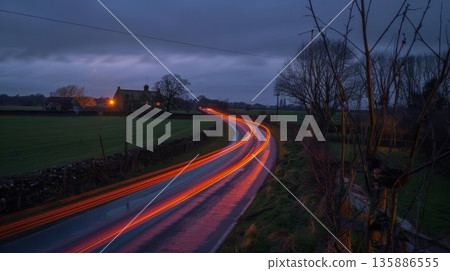 A peaceful countryside with a charming historic farm in the distance contrasted by the streaks of car light trails passing through the fields 135886555