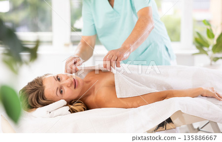 Relaxed middle-aged woman lying on massage table covered with blanket by masseuse Relaxed middle-aged woman lying on massage table covered with blanket by masseuse 135886667