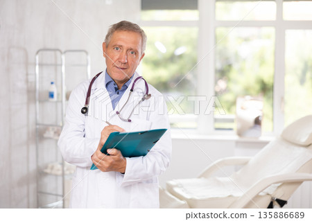 Old man doctor posing in doctor's cabinet with document in hands Old man doctor posing in doctor's cabinet with document in hands 135886689